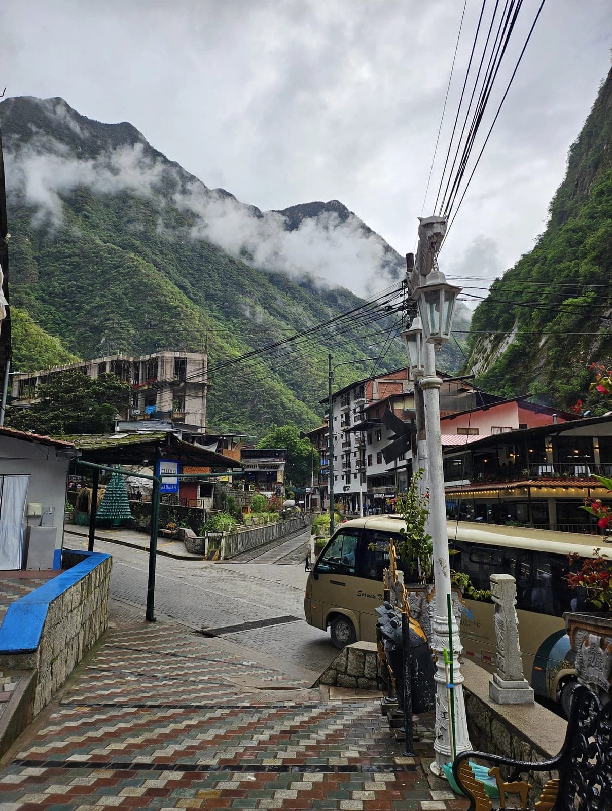 A street in Aguas Calientes (Machupicchu Pueblo) with a shuttle bus, below steep green mountains wrapped in cloud