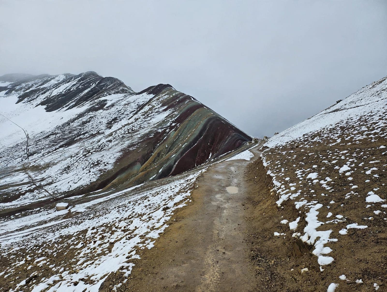 The striped slopes of Vinicunca (Rainbow Mountain) dusted with snow beside the trail, under heavy grey cloud