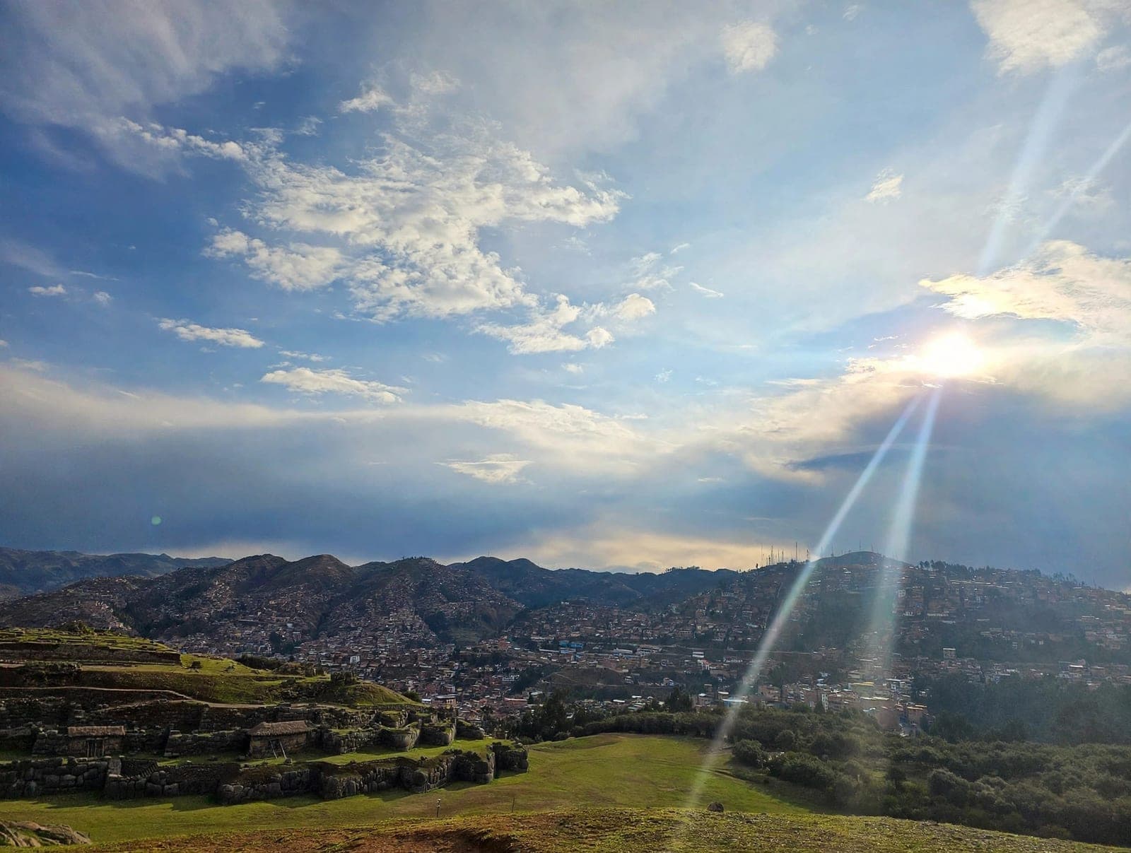 Cusco spread across its hills seen from Sacsayhuamán, with Inca stone terraces in the foreground and late sun breaking through cloud