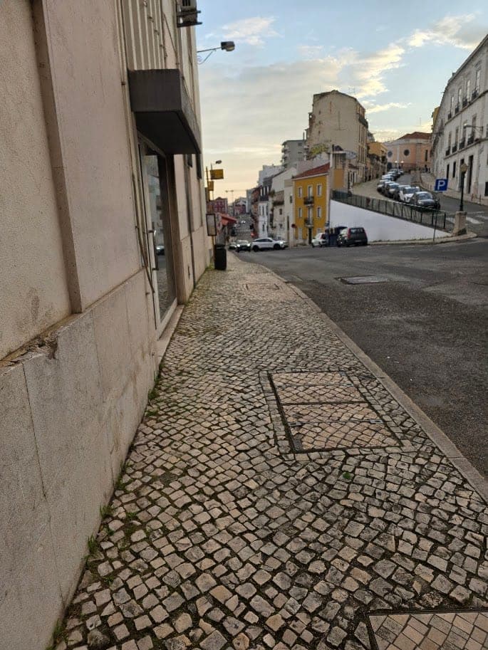 Une rue de Lisbonne avec un trottoir en petits pavés de pierre, des voitures stationnées et une route qui monte entre de vieux immeubles