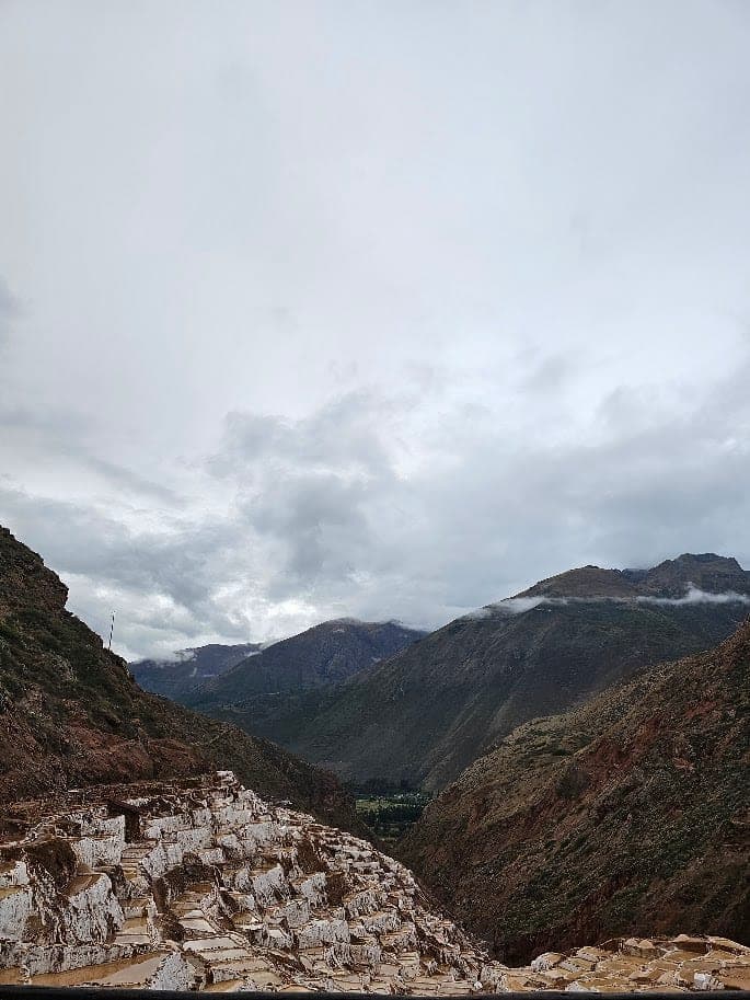 Terraced white salt pools of the Maras salt mines on a hillside, below cloud-topped mountains