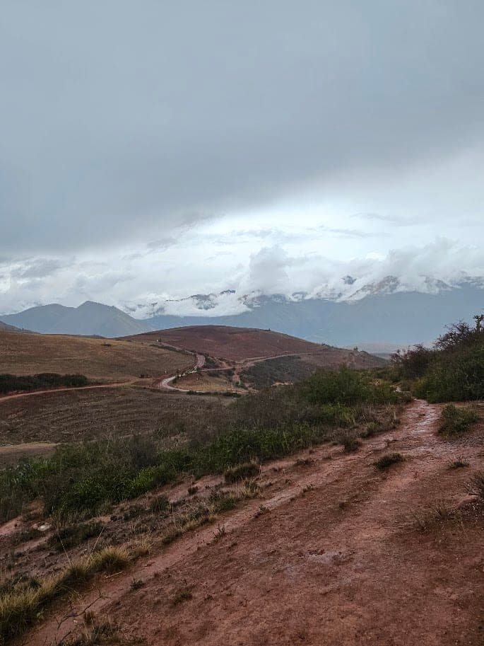 A red dirt trail through farmland in the Sacred Valley near Maras, with cloud hanging over snow-capped mountains